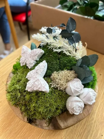 Jemná mechová dekorace s bílými růžemi, motýly a přírodními doplňky na dřevěném podkladu